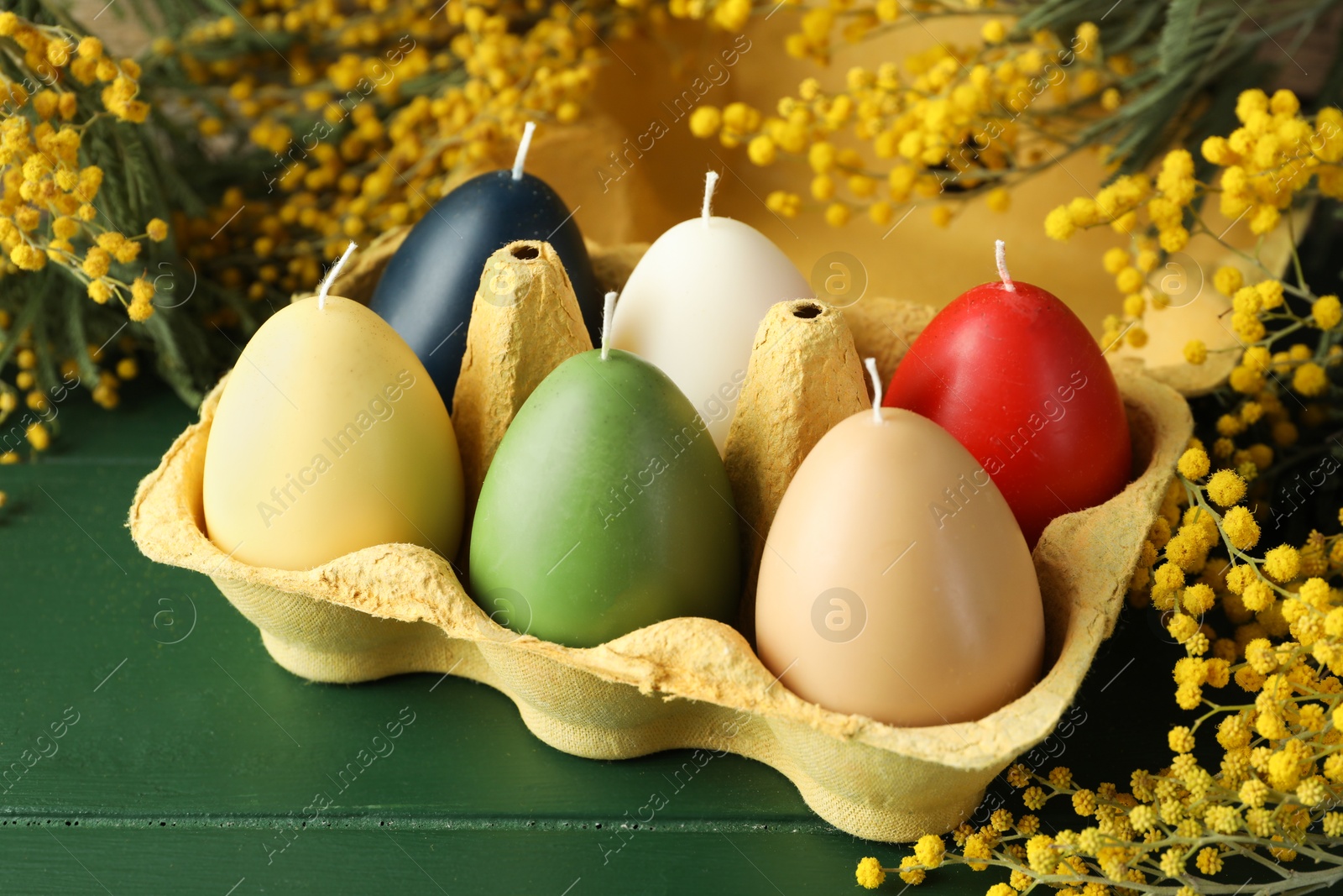 Photo of Colorful egg-shaped candles in carton and mimosa flowers on green wooden table, closeup. Easter decor