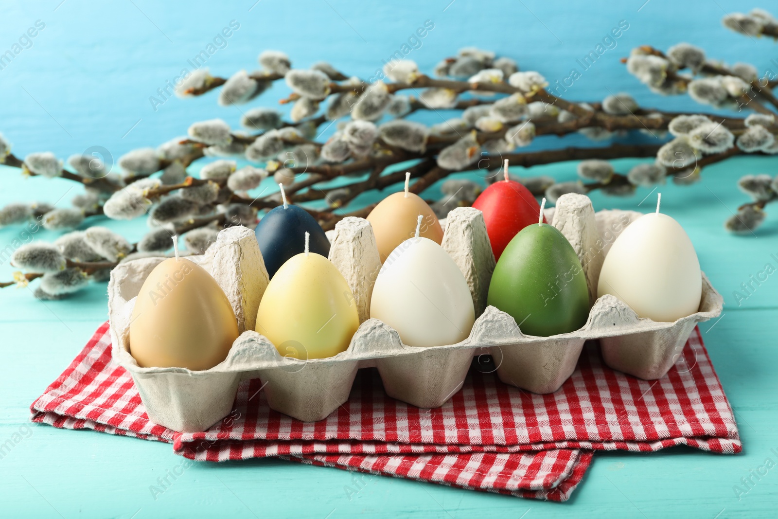 Photo of Colorful egg-shaped candles in carton and willow branches on turquoise table. Easter decor