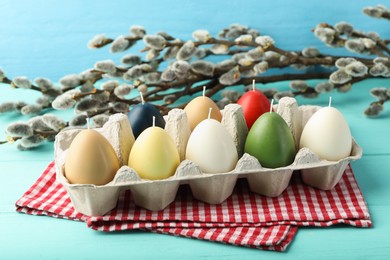 Photo of Colorful egg-shaped candles in carton and willow branches on turquoise table. Easter decor