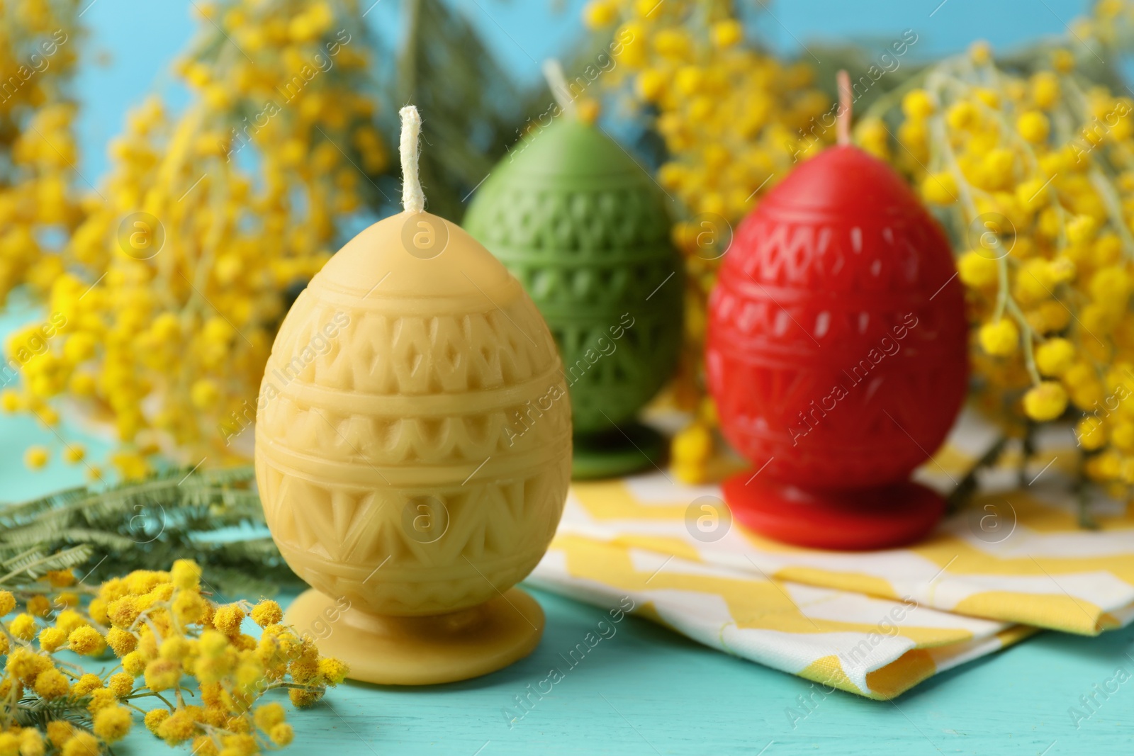 Photo of Colorful egg-shaped candles and mimosa flowers on turquoise wooden table, closeup. Easter decor