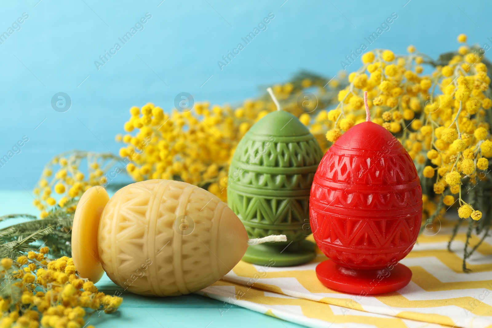 Photo of Colorful egg-shaped candles and mimosa flowers on turquoise table against light blue background, closeup. Easter decor