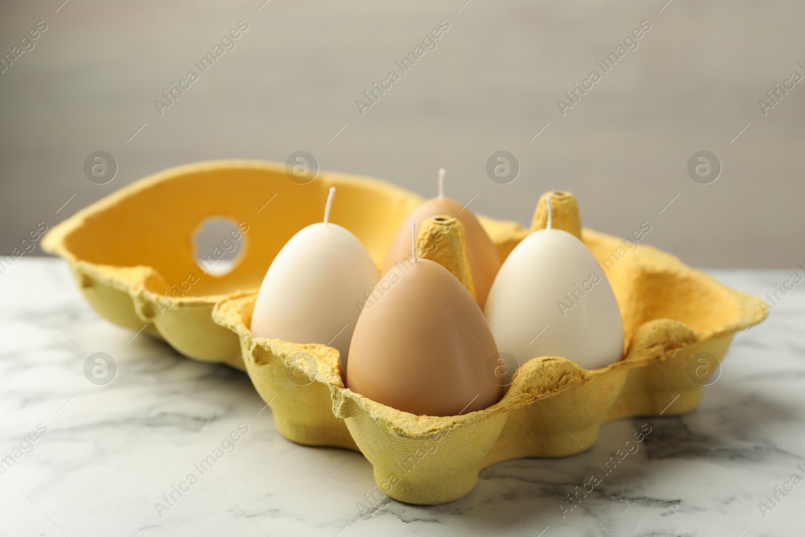 Photo of Egg-shaped candles in carton on white marble table against light gray background, closeup. Easter decor