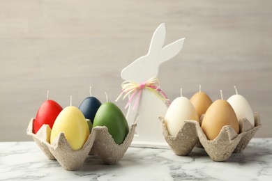 Photo of Colorful egg-shaped candles in carton and bunny figure on white marble table against light gray background. Easter decor