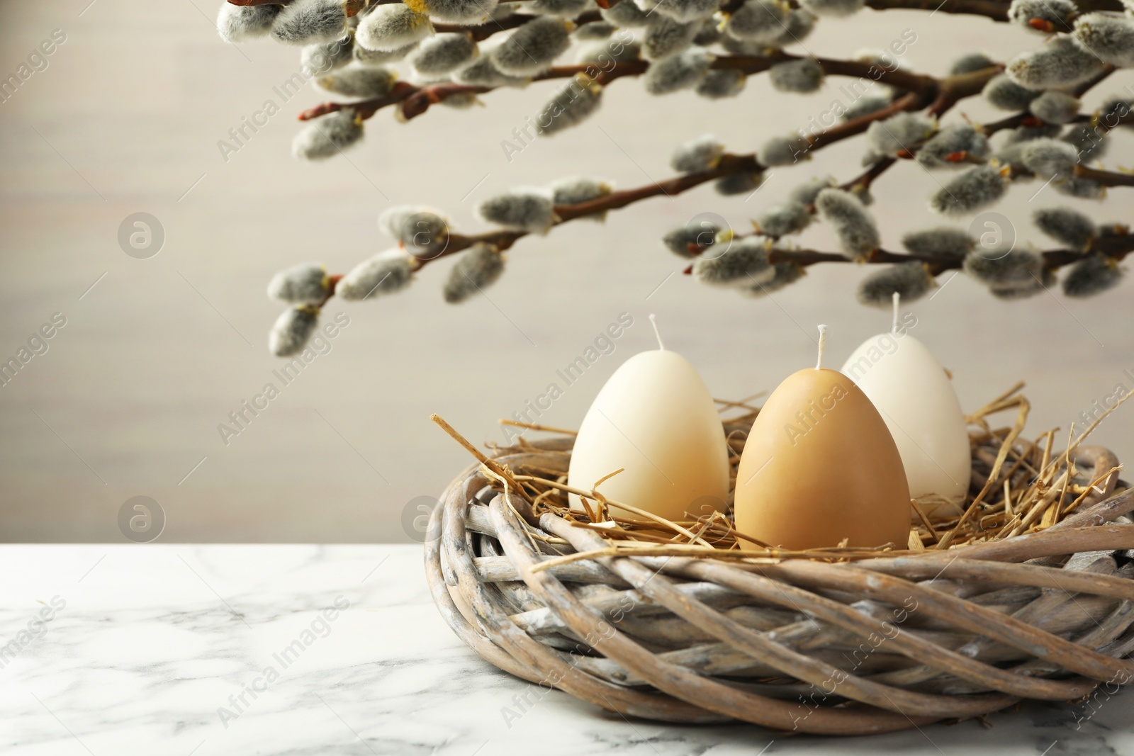 Photo of Egg-shaped candles in nest on white marble table and willow branches against light background, space for text. Easter decor