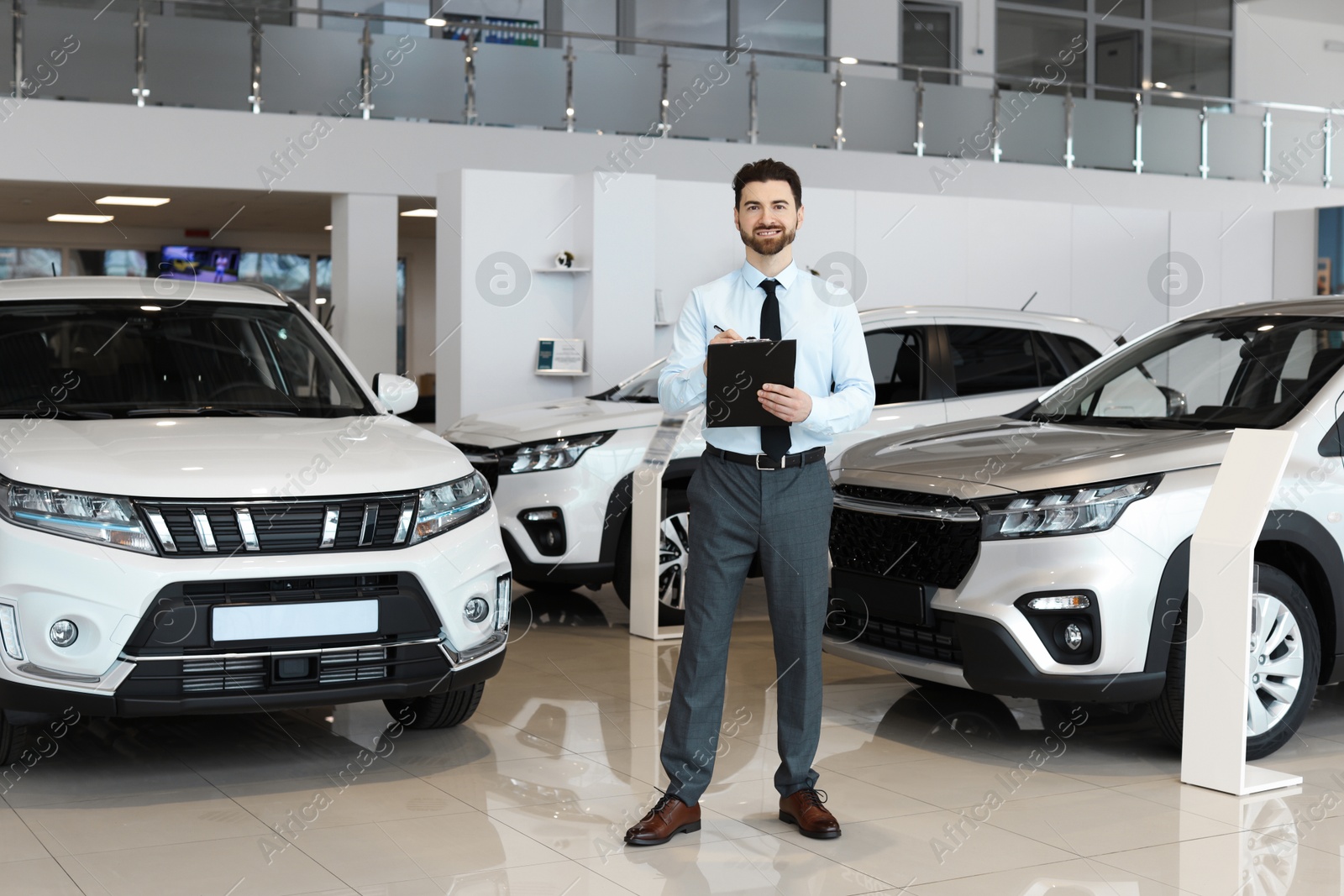 Photo of Happy salesman with clipboard near new cars in salon