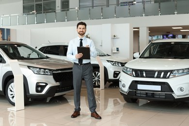 Photo of Happy salesman with clipboard near new cars in salon