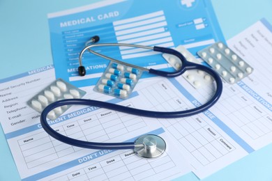 Photo of Stethoscope, medical card forms and pills on light blue background, closeup