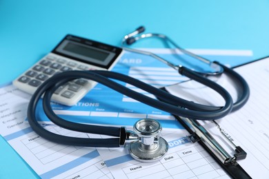 Photo of Stethoscope, calculator and medical card forms on light blue background, closeup