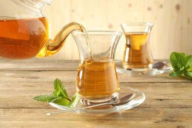 Photo of Pouring freshly brewed tea from teapot into glass cup on wooden table, closeup