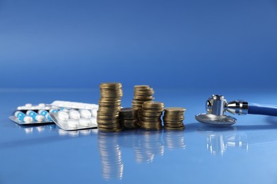 Photo of Stethoscope, pills and coins on glass surface against blue background
