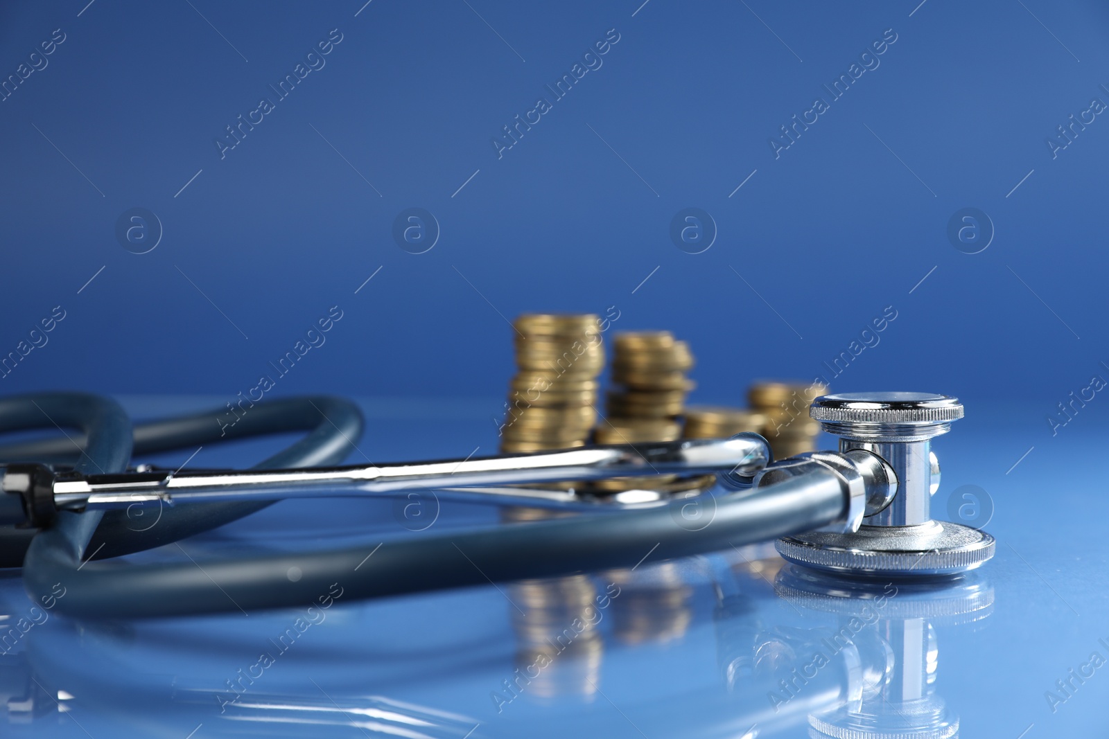 Stethoscope and coins on glass surface against blue background, closeup Photo of Stethoscope and coins on glass surface against blue background, closeup