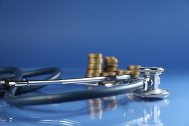 Photo of Stethoscope and coins on glass surface against blue background, closeup
