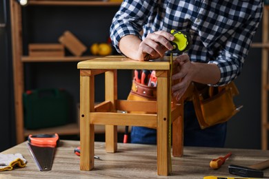 Photo of Repairman measuring wooden stool at table in workshop, closeup