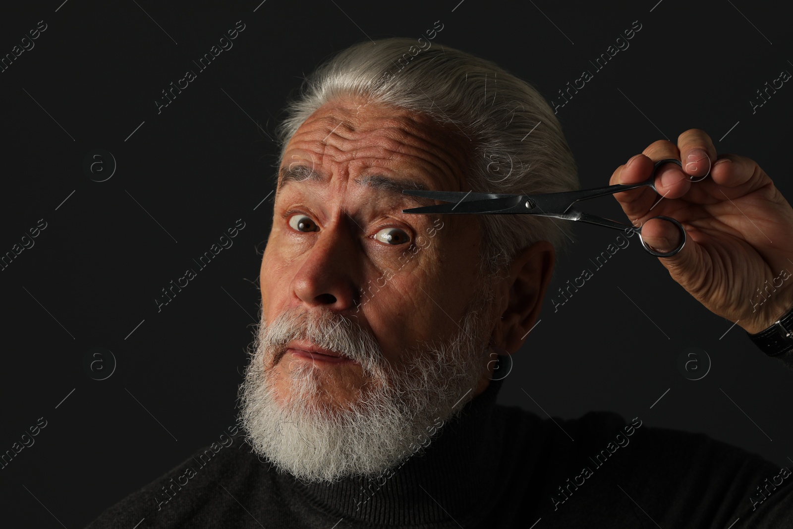 Photo of Bearded senior man trimming eyebrow with scissors on dark grey background