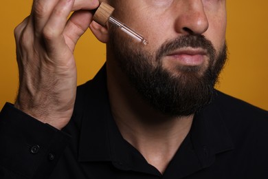 Photo of Man applying serum onto his beard on orange background, closeup