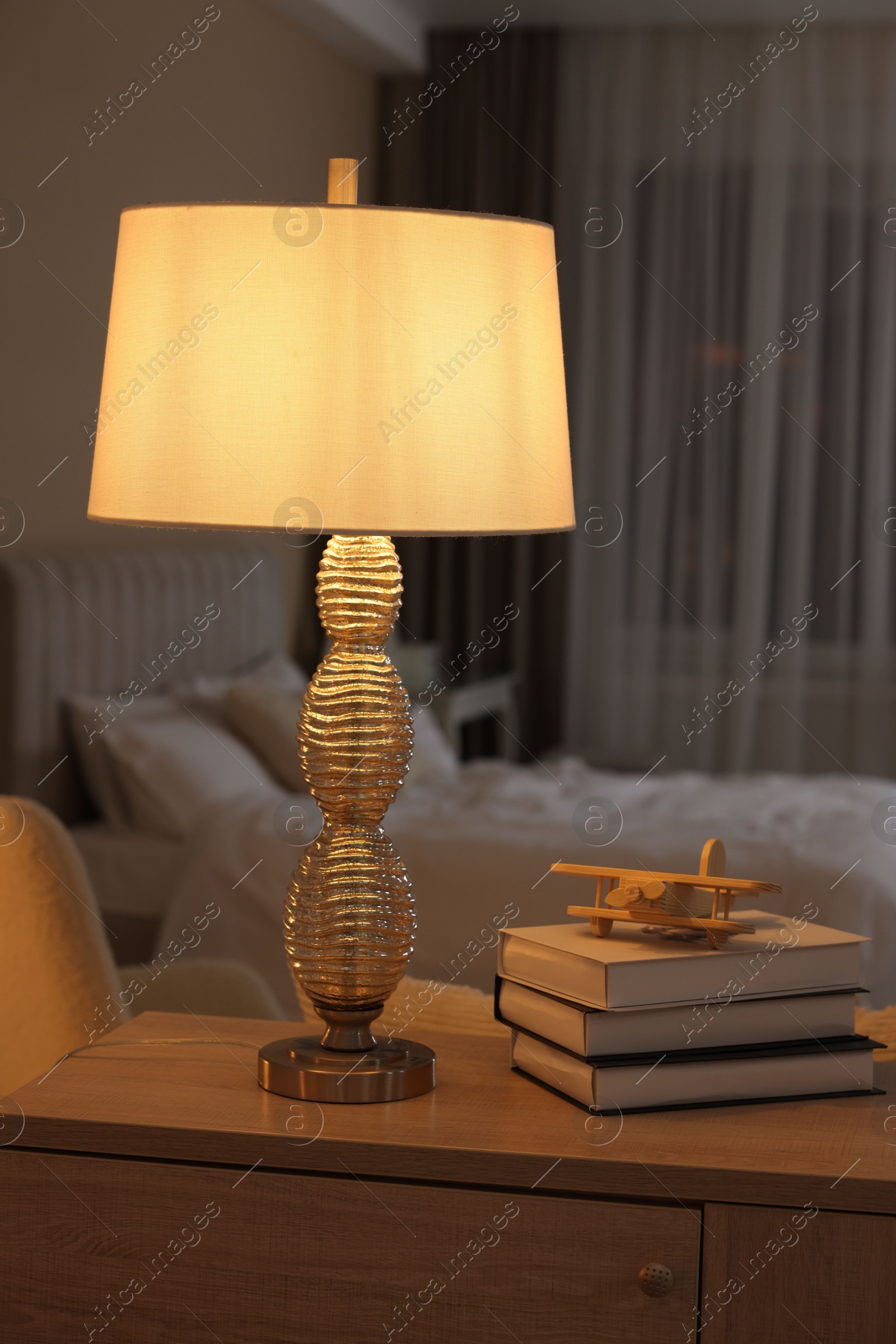 Photo of Beautiful night lamp, stack of books and airplane model on chest of drawers in bedroom