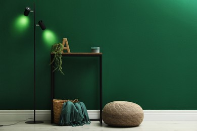 Photo of Stylish knitted pouf, lamp and table near green wall indoors, space for text