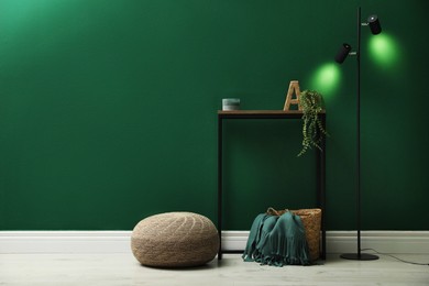 Photo of Stylish knitted pouf, lamp and table near green wall indoors, space for text