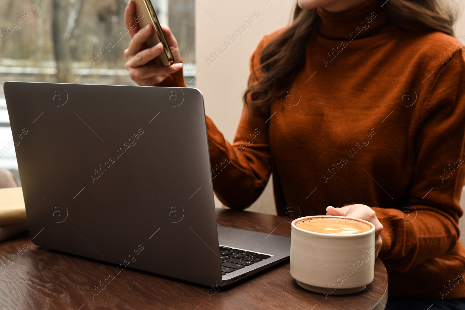 Photo of Woman with smartphone and laptop having coffee break at wooden table in cafe, closeup