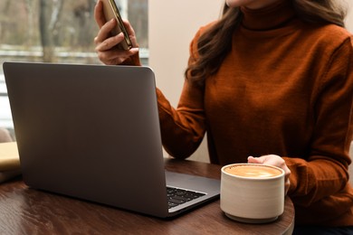 Photo of Woman with smartphone and laptop having coffee break at wooden table in cafe, closeup