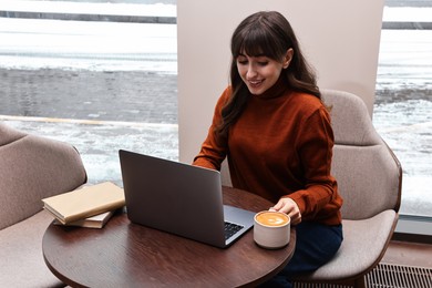 Smiling woman with laptop having coffee break at wooden table in cafe