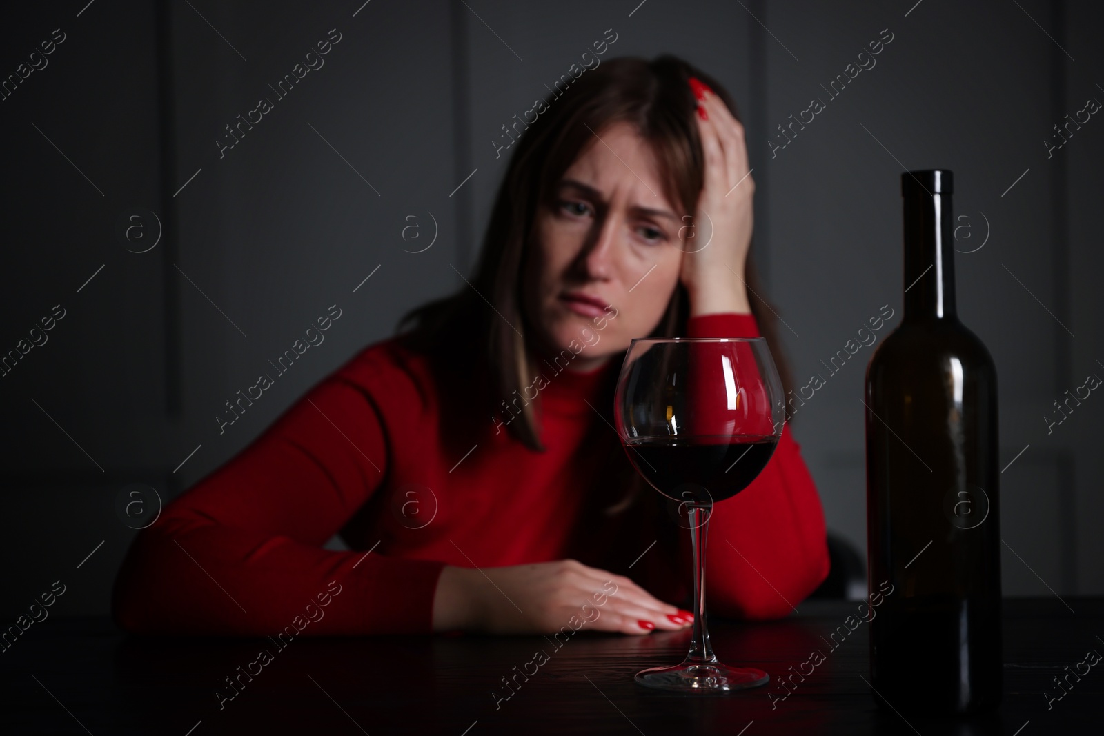 Photo of Alcohol addiction. Woman at wooden table indoors, focus on glass of red wine and bottle