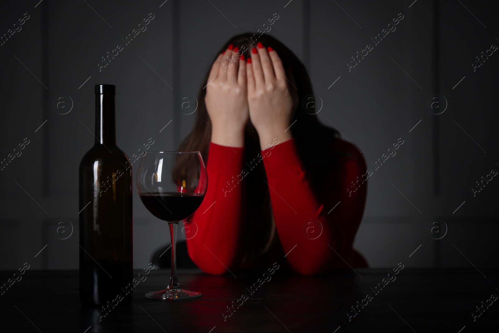 Photo of Alcohol addiction. Woman covering her face at wooden table indoors, focus on glass of red wine and bottle