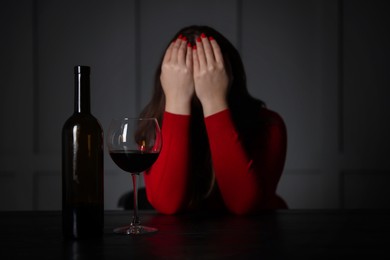 Photo of Alcohol addiction. Woman covering her face at wooden table indoors, focus on glass of red wine and bottle