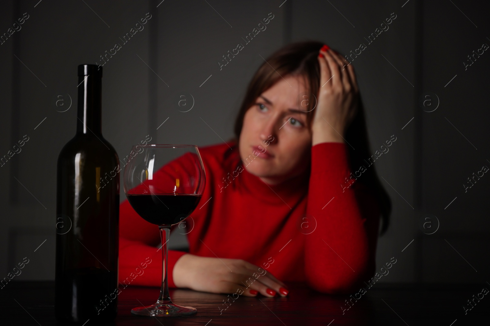 Photo of Alcohol addiction. Woman at wooden table indoors, focus on glass of red wine and bottle