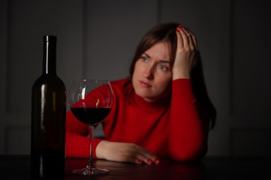 Photo of Alcohol addiction. Woman at wooden table indoors, focus on glass of red wine and bottle