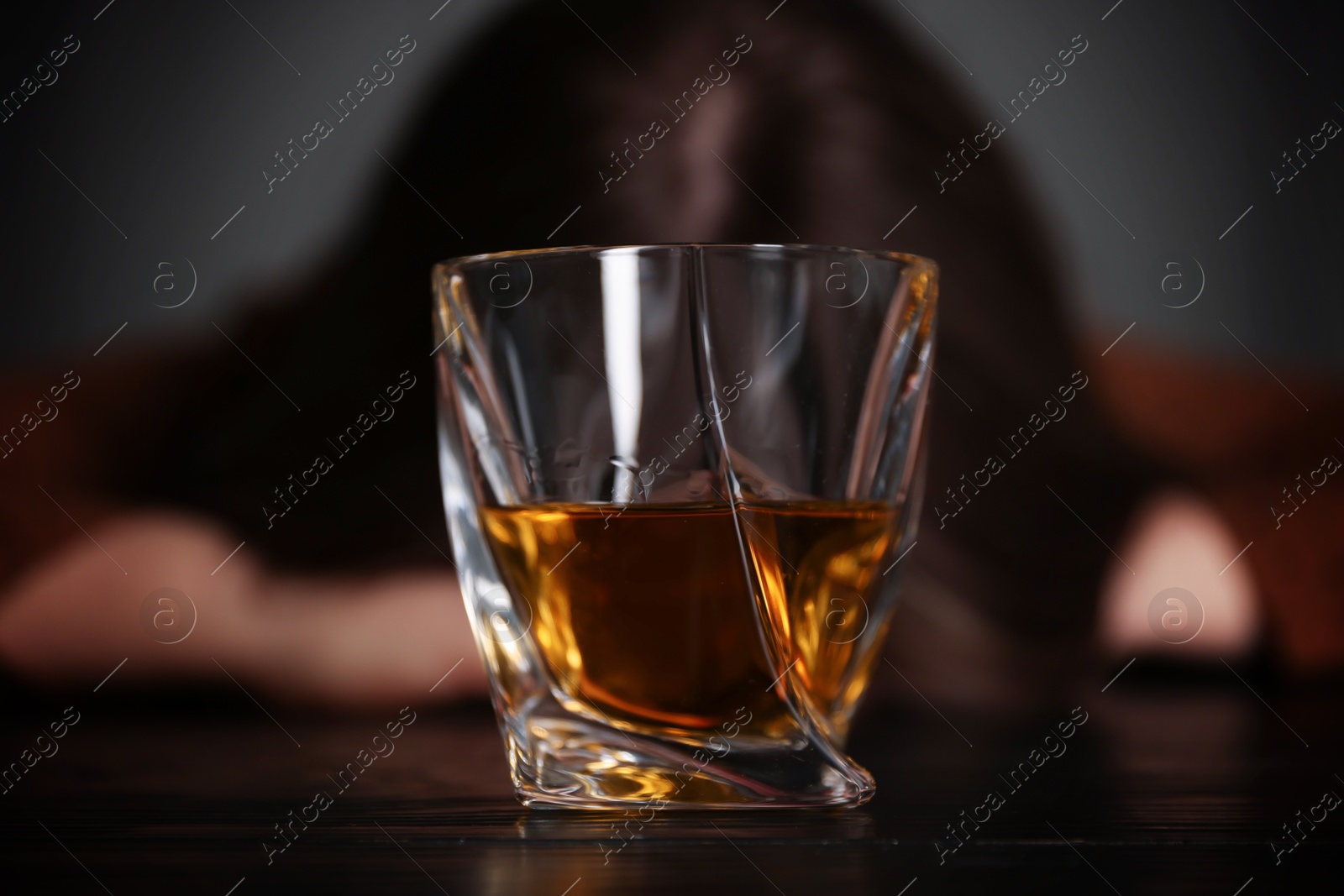 Photo of Alcohol addiction. Woman at wooden table indoors, focus on glass of whiskey