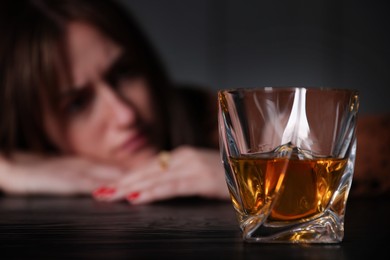 Photo of Alcohol addiction. Woman at wooden table indoors, focus on glass of whiskey