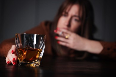 Photo of Alcohol addiction. Woman with glass of whiskey smoking cigarette at table indoors, selective focus