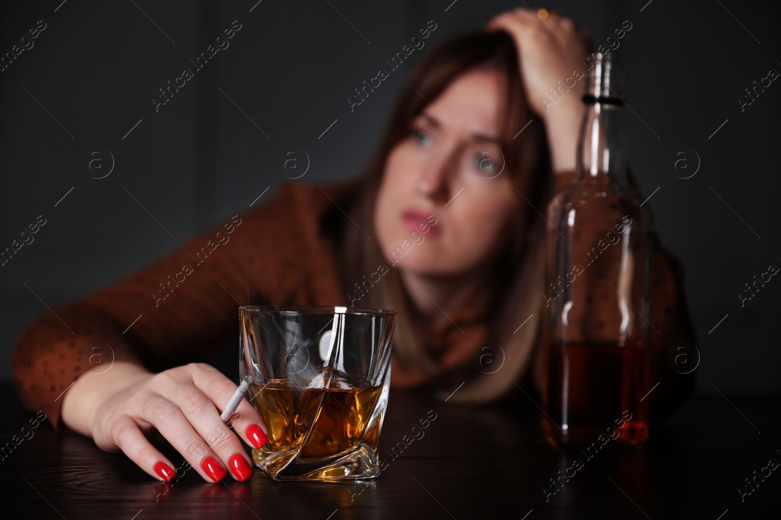 Photo of Alcohol addiction. Woman with glass of whiskey, cigarette and bottle at table indoors, selective focus