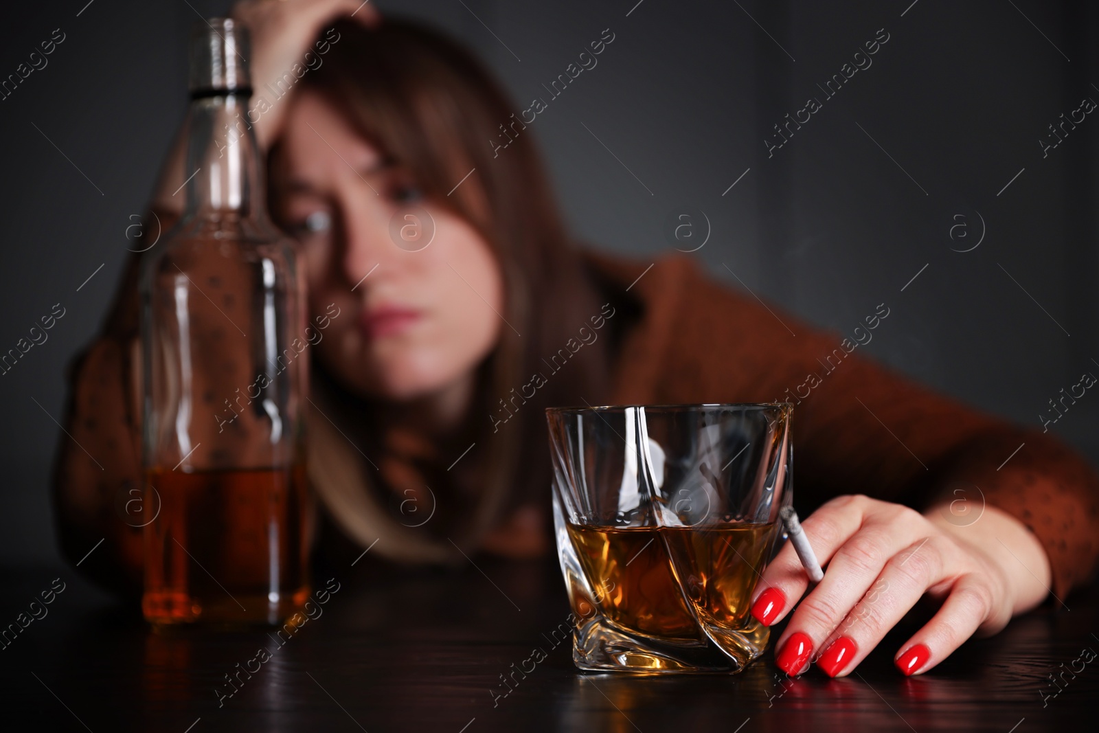 Photo of Alcohol addiction. Woman with glass of whiskey, cigarette and bottle at table indoors, selective focus