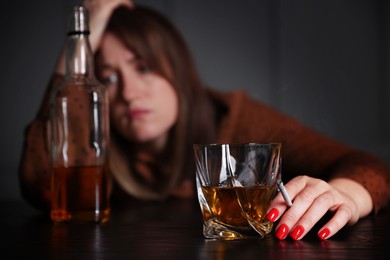 Photo of Alcohol addiction. Woman with glass of whiskey, cigarette and bottle at table indoors, selective focus