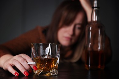 Photo of Alcohol addiction. Woman with glass of whiskey, cigarette and bottle at table indoors, selective focus