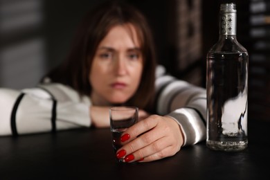 Photo of Alcohol addiction. Woman with shot glass of vodka and bottle at table indoors, selective focus