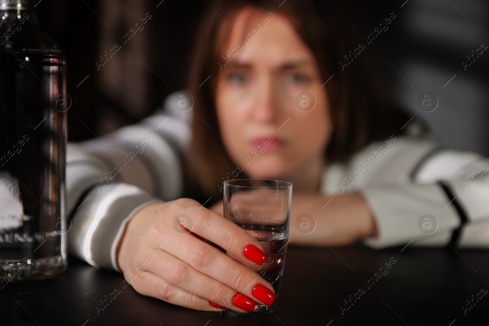 Photo of Alcohol addiction. Woman with shot glass of vodka and bottle at table indoors, selective focus