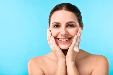 Photo of Smiling woman washing her face with cleansing foam against light blue background, space for text