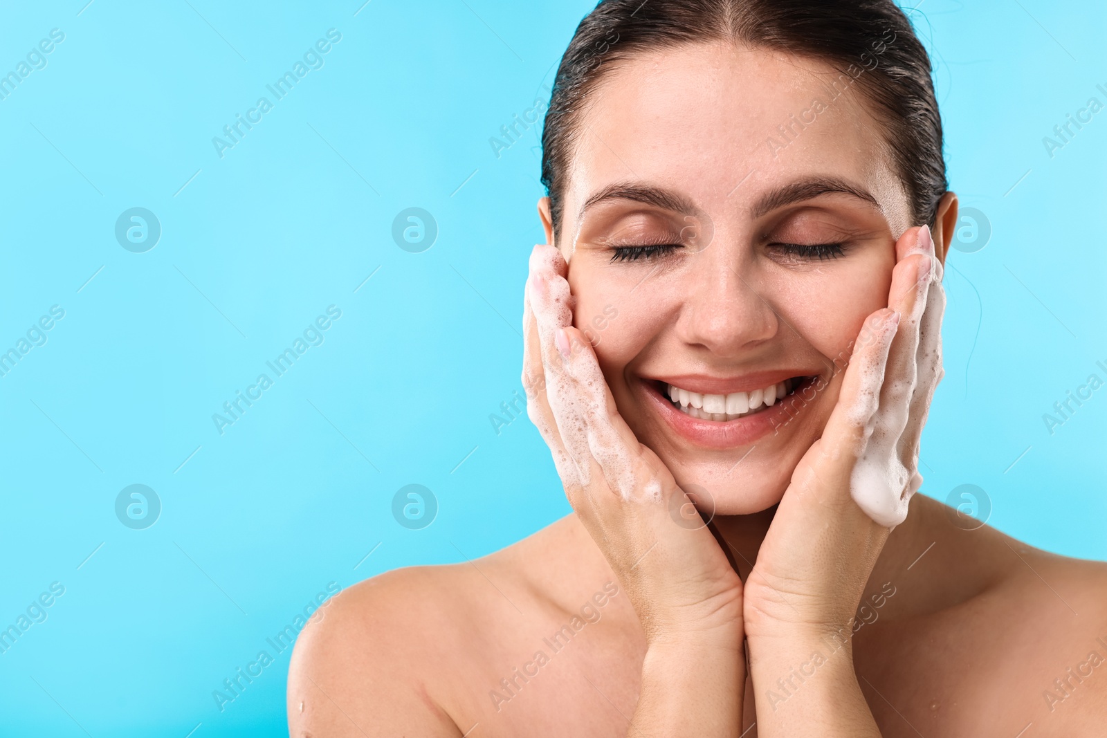 Photo of Smiling woman washing her face with cleansing foam against light blue background, space for text