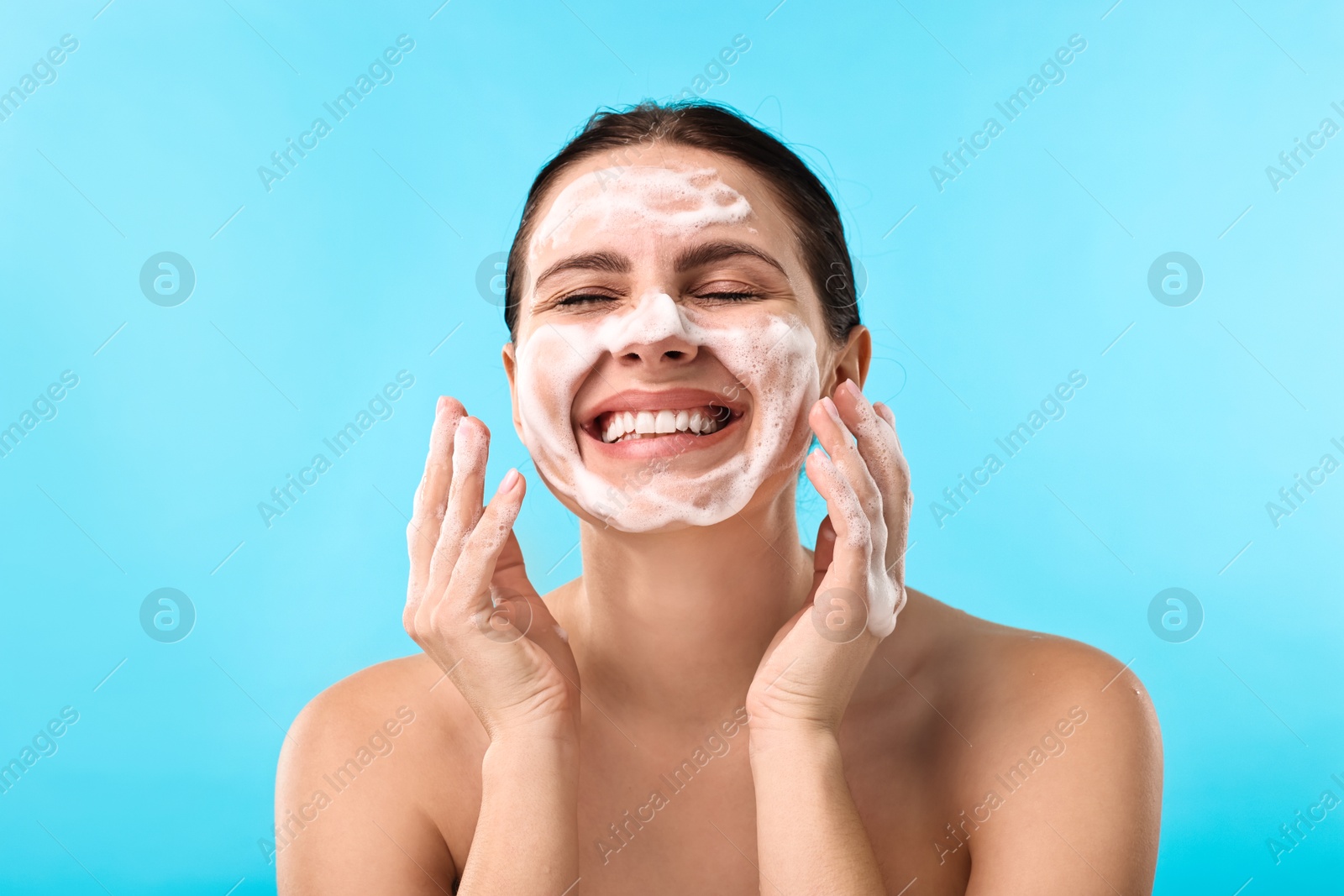 Photo of Smiling woman washing her face with cleansing foam on light blue background