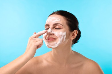 Photo of Cute woman with cleansing foam on her face against light blue background. Washing routine