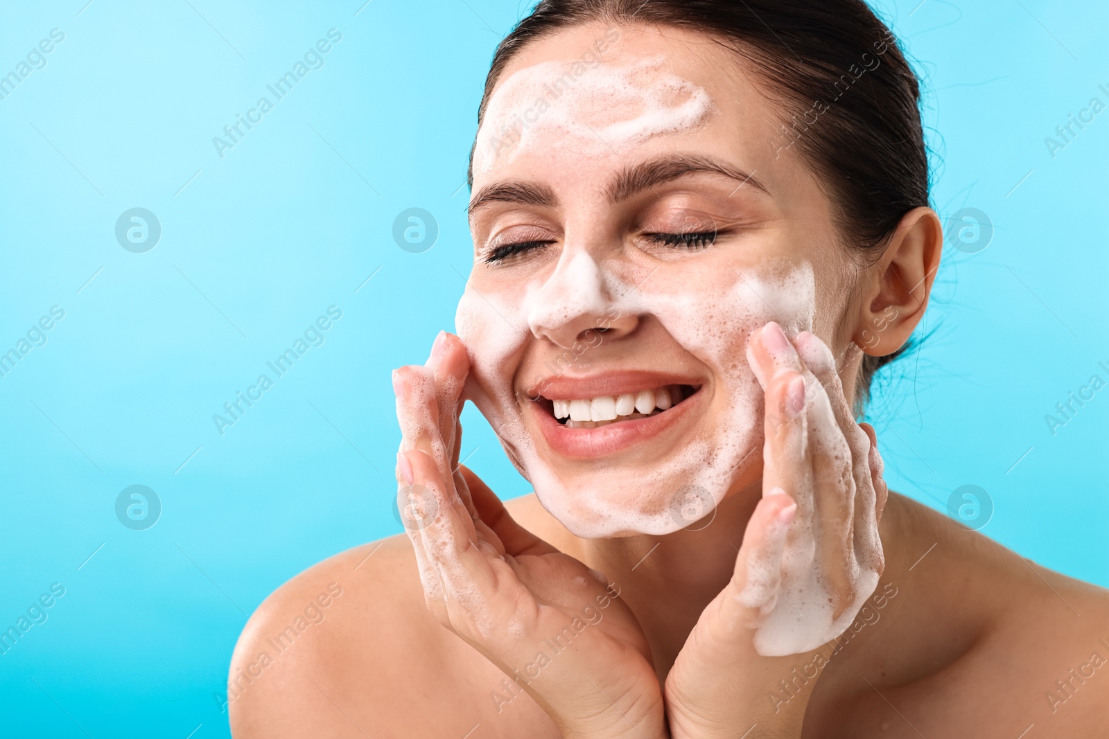 Photo of Smiling woman washing her face with cleansing foam on light blue background, space for text