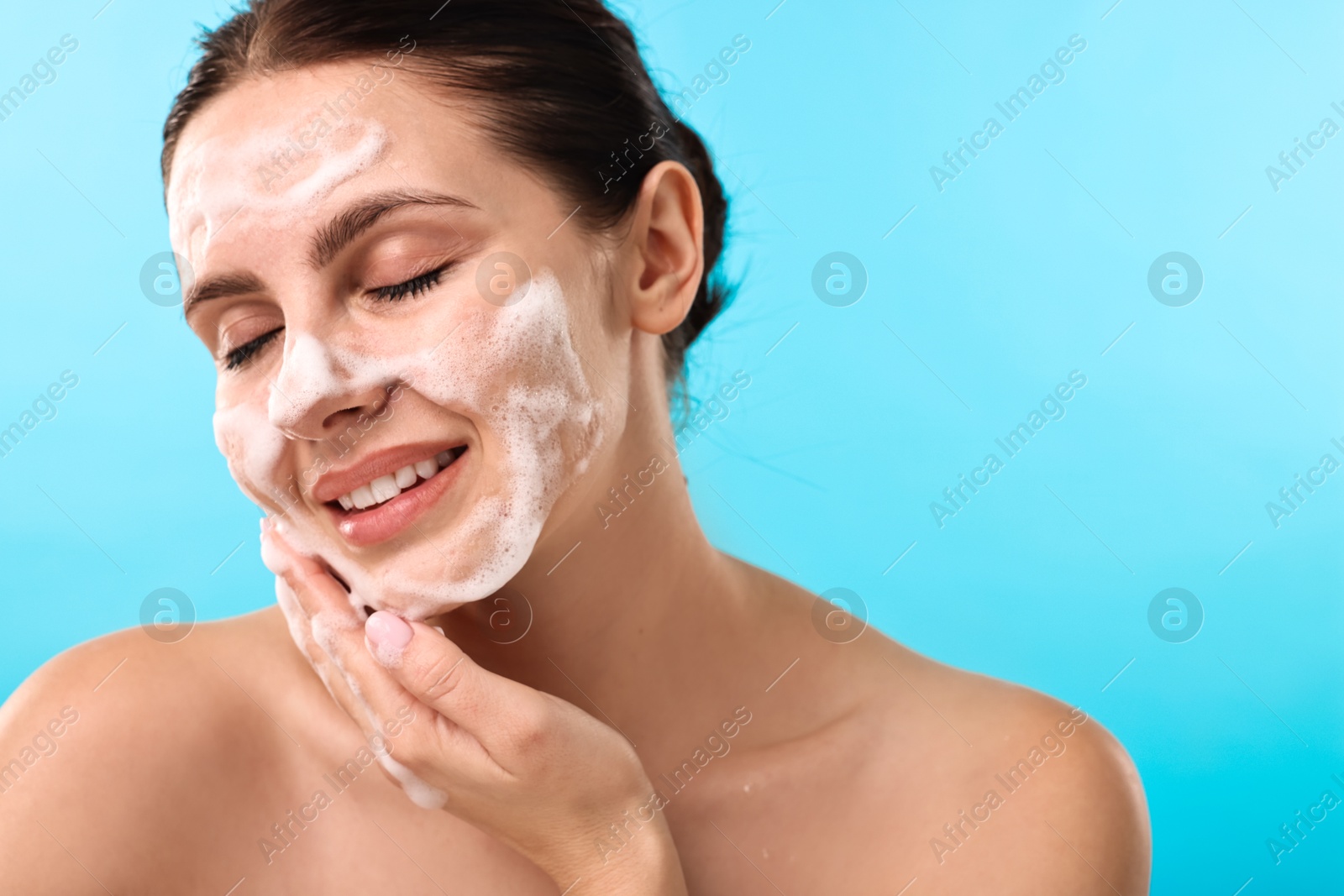 Photo of Smiling woman washing her face with cleansing foam on light blue background, space for text