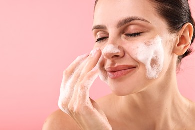 Photo of Attractive woman washing her face with cleansing foam on pink background, space for text