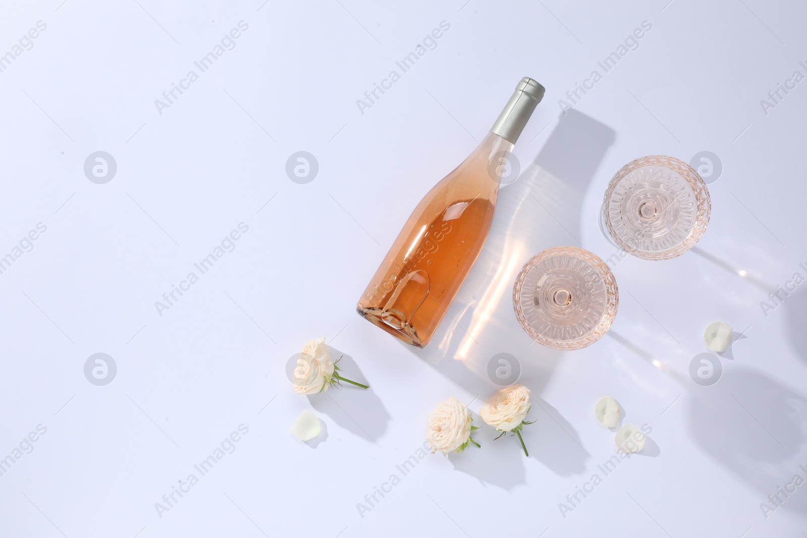 Photo of Delicious rose wine in glasses and bottle with flowers on white background, flat lay. Space for text