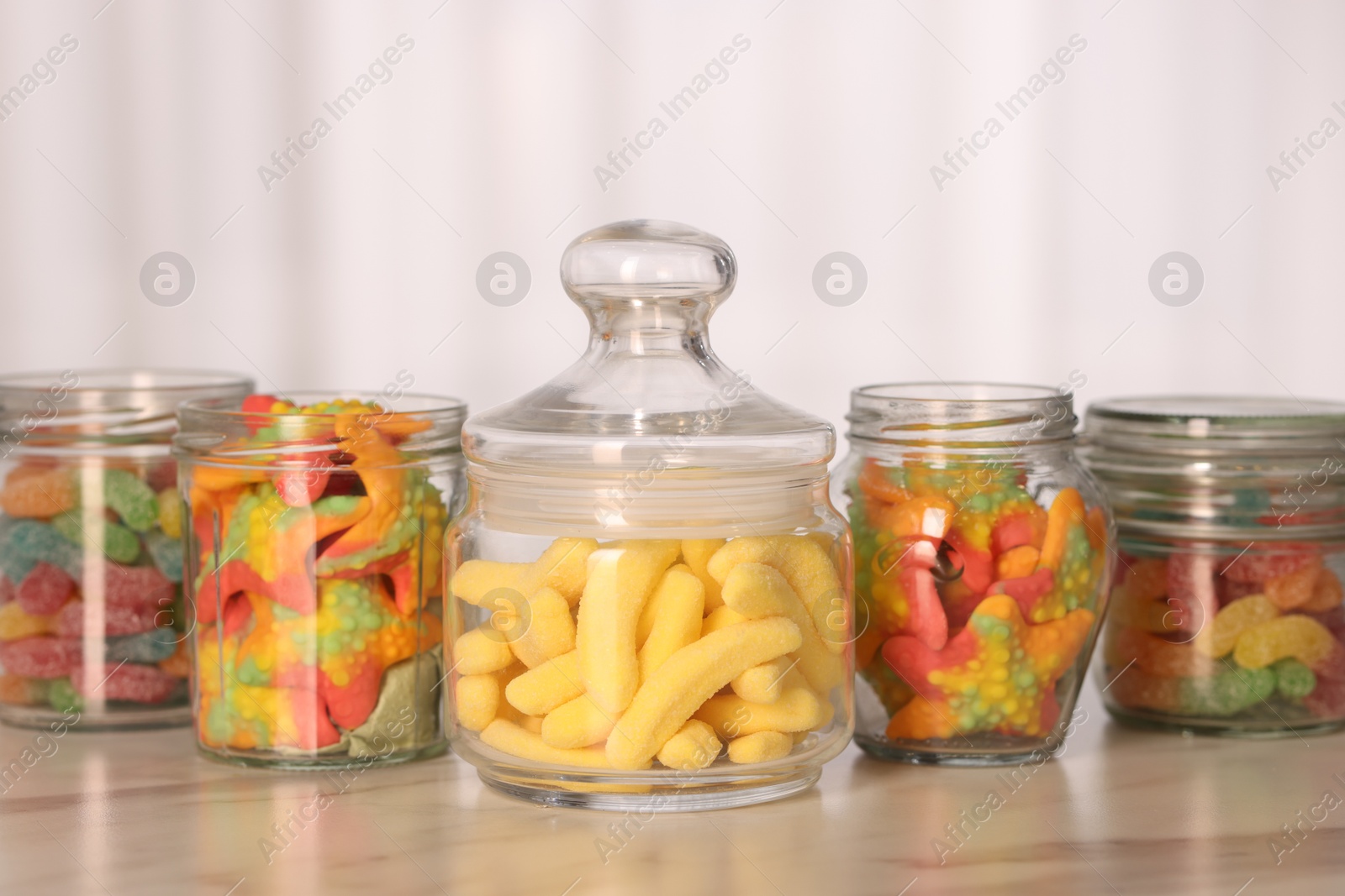 Photo of Tasty gummy candies in glass jars on white marble table, closeup