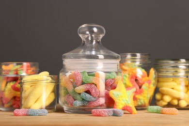 Photo of Tasty gummy candies in glass jars on wooden table against grey background, closeup