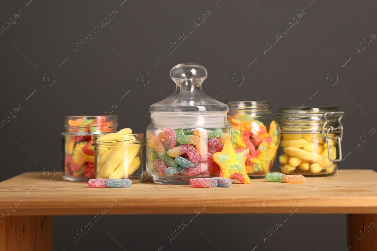 Photo of Tasty gummy candies in glass jars on wooden table against grey background